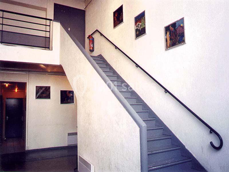 Escalier intérieur en métal gris avec une rampe noire, entouré de murs blancs décorés de tableaux encadrés.