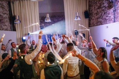 Des invités dansent et agitent des bâtons lumineux dans une salle décorée avec des murs en pierre et des rideaux clairs.