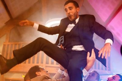 Un homme en costume est soulevé dans les airs par plusieurs personnes lors d'une fête dans une salle en bois.