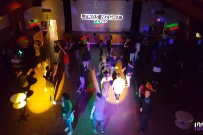 Des personnes dansent dans une salle sombre éclairée par des lumières colorées, avec un écran affichant ’Linas Night Fever’.