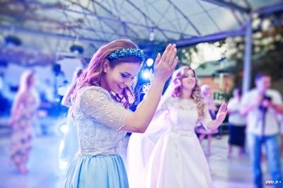 Deux femmes en robes élégantes dansent sous une structure couverte, entourées de lumière et d'autres personnes.