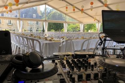 Salle de réception sous une tente avec tables dressées, vue depuis une table de DJ équipée de matériel audio.
