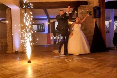 Un couple en tenue de mariage entre dans une salle décorée, entouré d'effets pyrotechniques au sol.