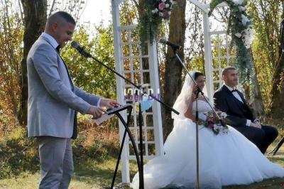 Un homme en costume parle au micro lors d'une cérémonie de mariage en plein air, avec les mariés assis à l'arrière-plan.