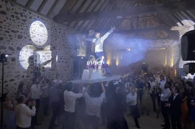 Un homme debout sur une table portée par des invités dans une salle en pierre lors d'une fête animée.