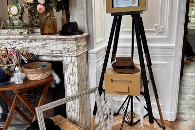 Borne photo sur trépied dans un salon élégant avec cheminée, fleurs, accessoires et cadre décoratif au sol.