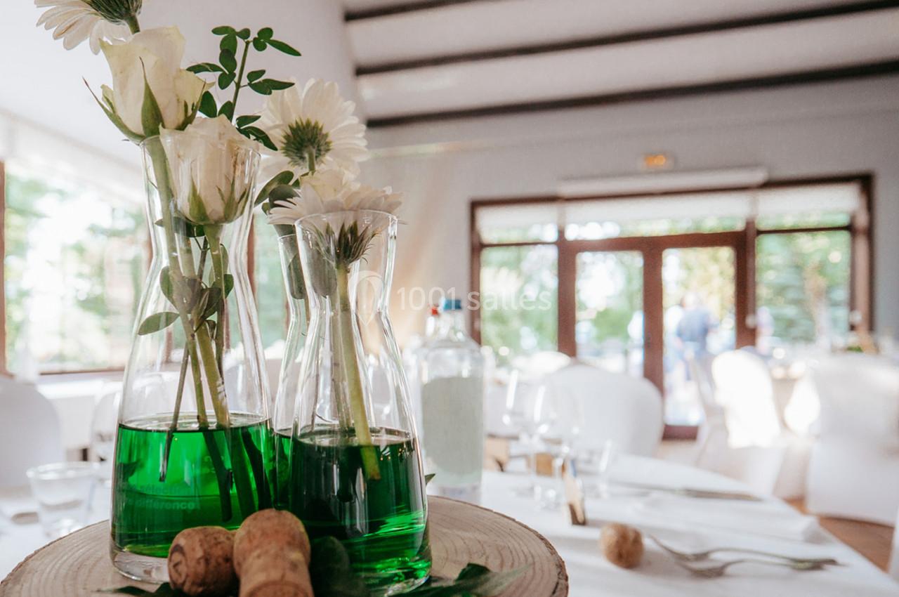 Vases en verre contenant des fleurs blanches et un liquide vert, posés sur une rondelle de bois dans une salle lumineuse.