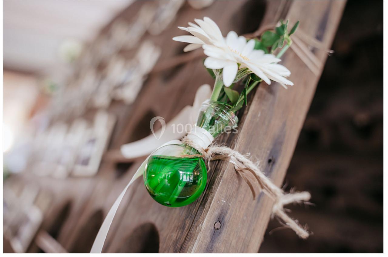Petit vase en verre vert contenant une fleur blanche, attaché à une structure en bois avec une corde.