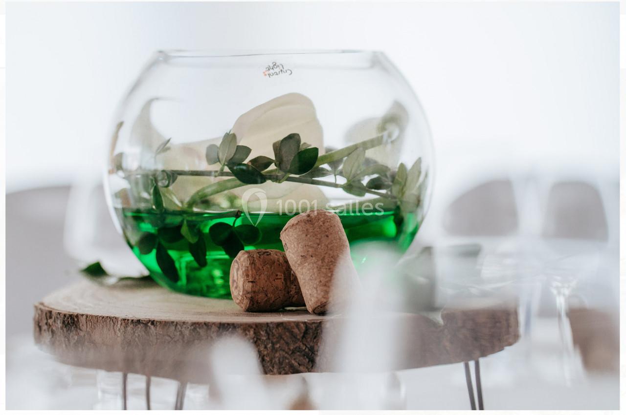 Vase rond avec liquide vert, feuillage et fleurs blanches, posé sur une tranche de bois avec deux bouchons en liège.