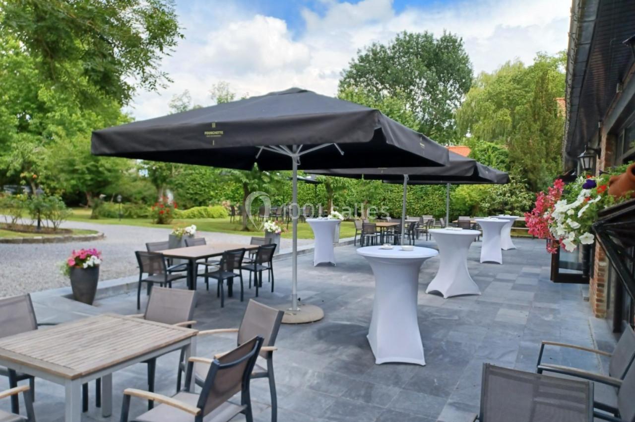Terrasse extérieure avec tables, chaises, parasols noirs et tables hautes, entourée de verdure et fleurs.