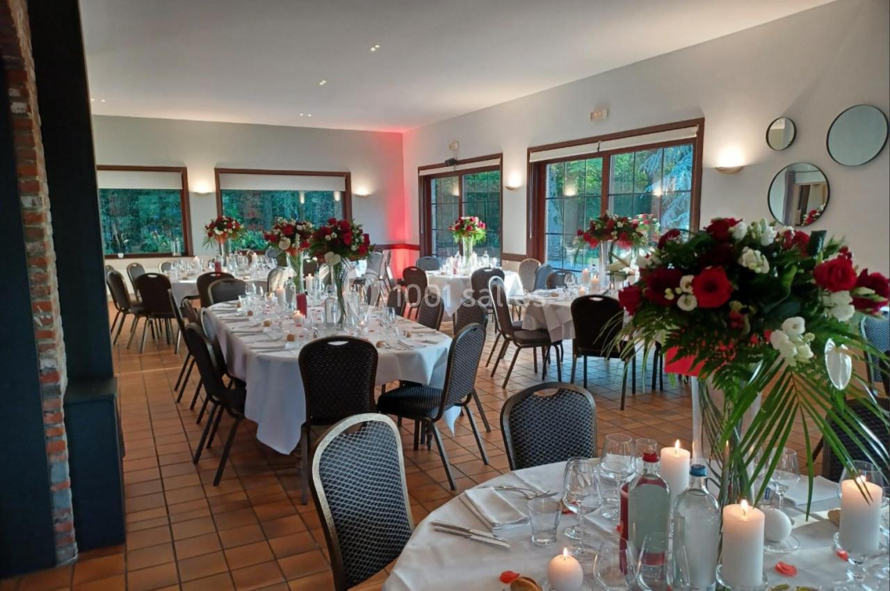 Salle de réception décorée avec des tables rondes dressées, ornées de bouquets de fleurs rouges et blanches.