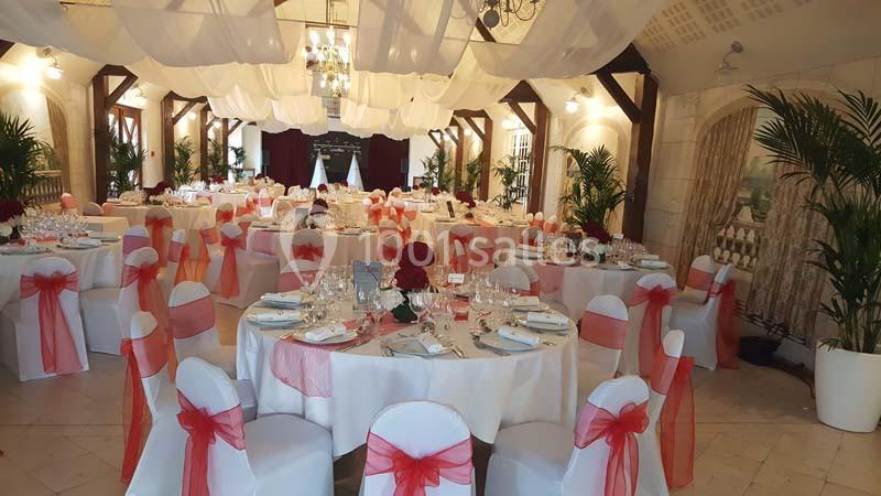 Salle de réception décorée avec des tables rondes, nappes blanches, nœuds rouges sur les chaises et tentures suspendues.