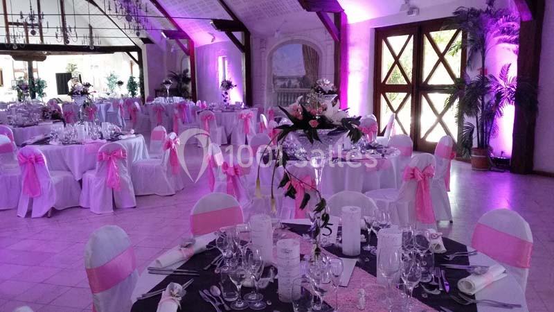 Salle de réception décorée avec des tables rondes, nappes blanches, rubans roses et éclairage d'ambiance violet.