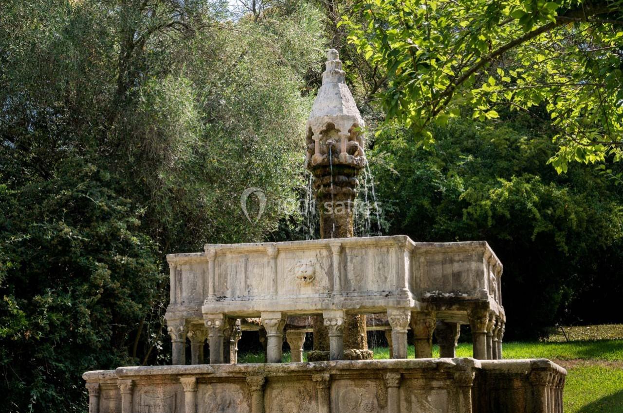 Fontaine en pierre ancienne entourée de végétation dense, avec des jets d'eau coulant depuis plusieurs niveaux.