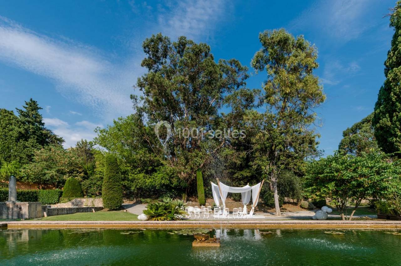 Un jardin avec un bassin, des arbres et une structure blanche décorative entourée de chaises.