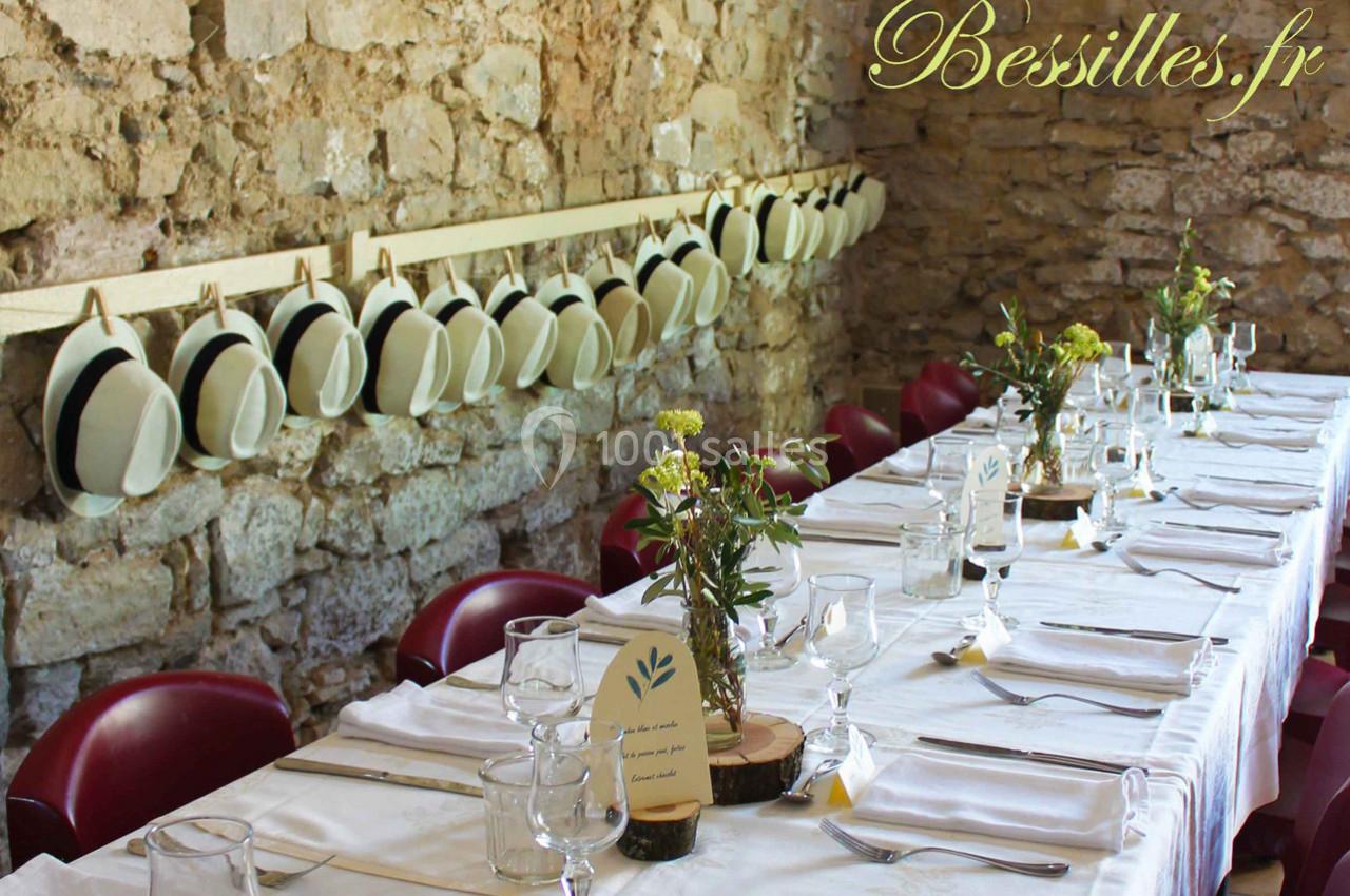 Table dressée avec nappes blanches, décor champêtre, chapeaux suspendus sur un mur en pierre.