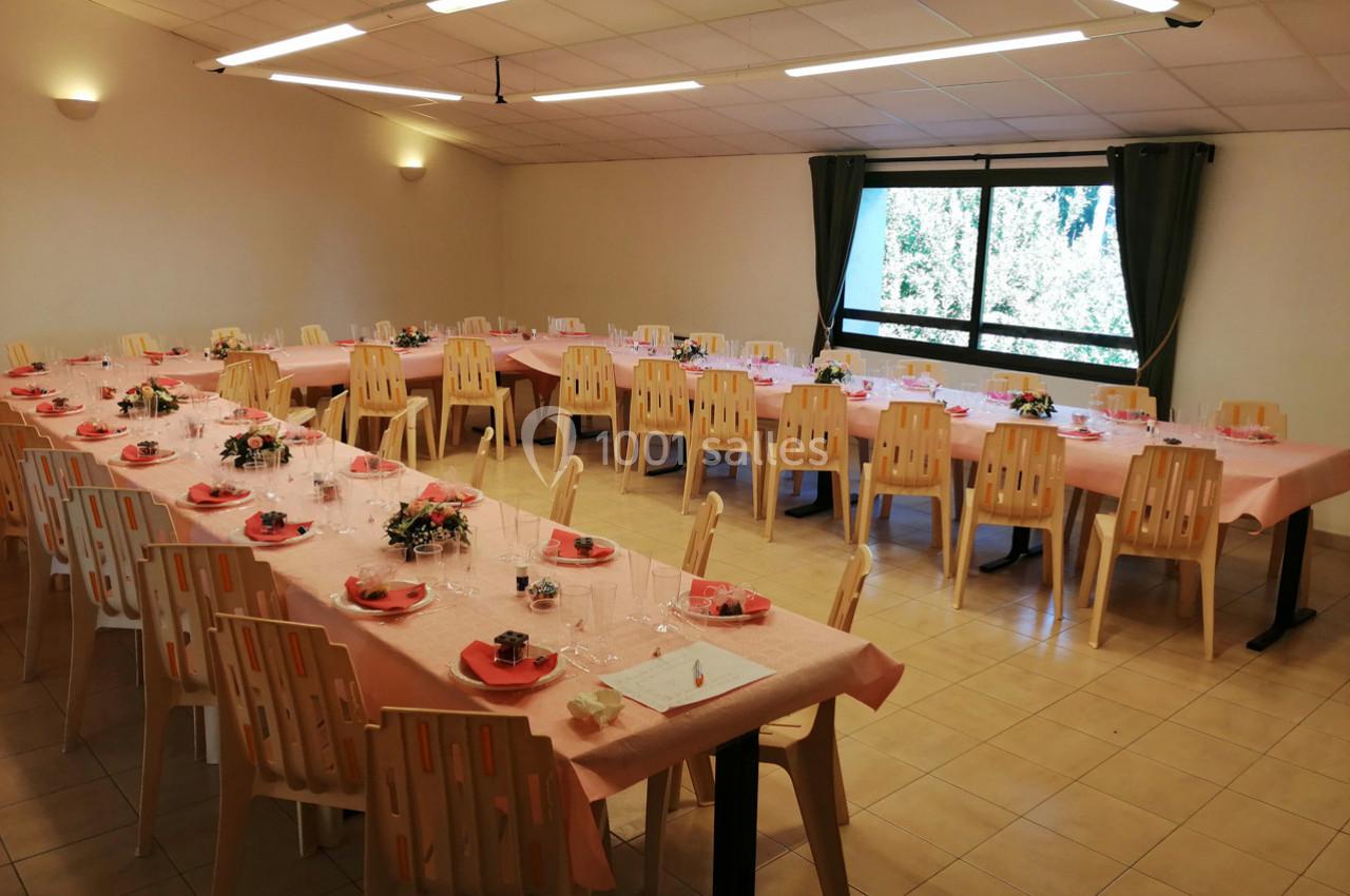 Salle décorée pour un repas, avec tables alignées, nappes roses, chaises en plastique et fenêtres donnant sur un extérieur…