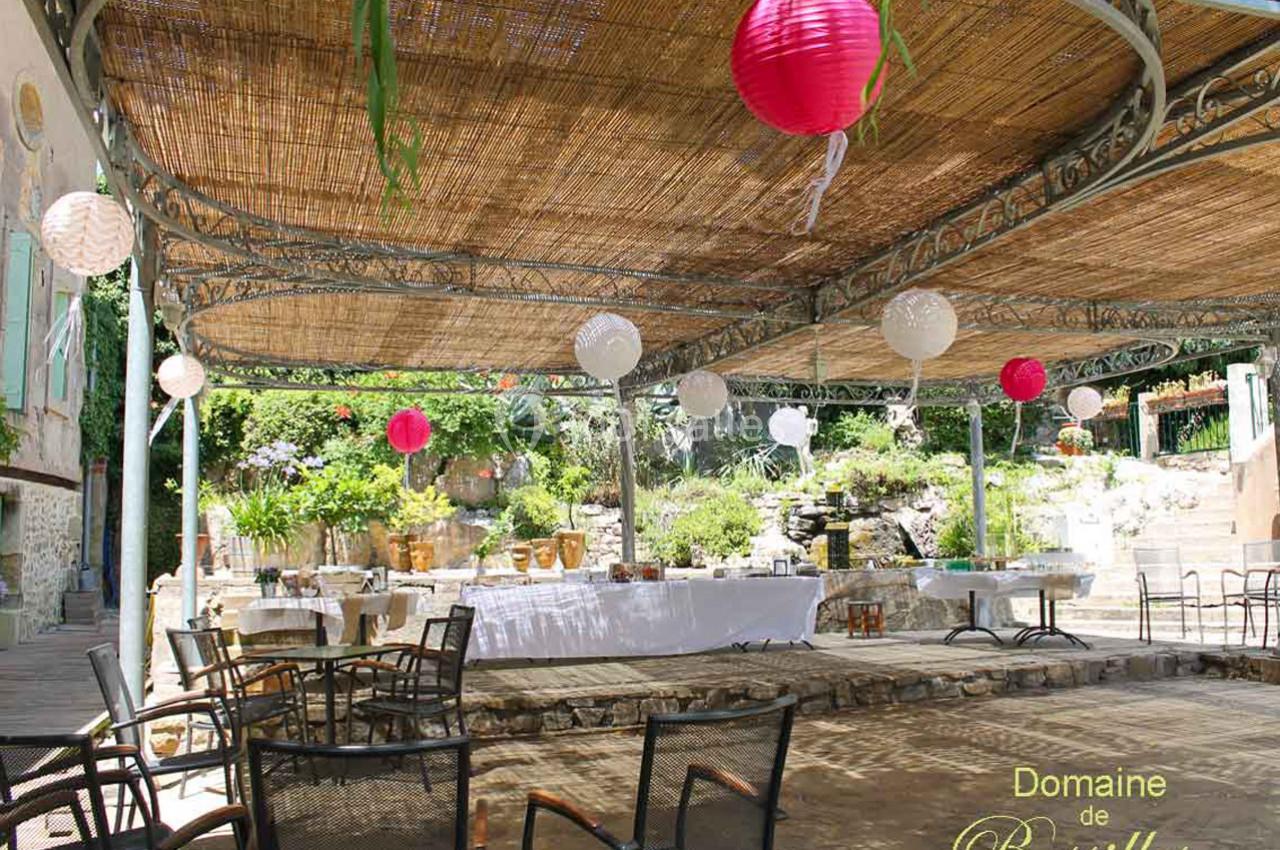 Terrasse ombragée avec tables dressées, lanternes suspendues et végétation en arrière-plan.