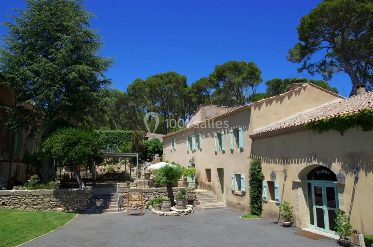 Cour d'une maison en pierre avec volets verts, entourée d'arbres et d'un ciel bleu dégagé.