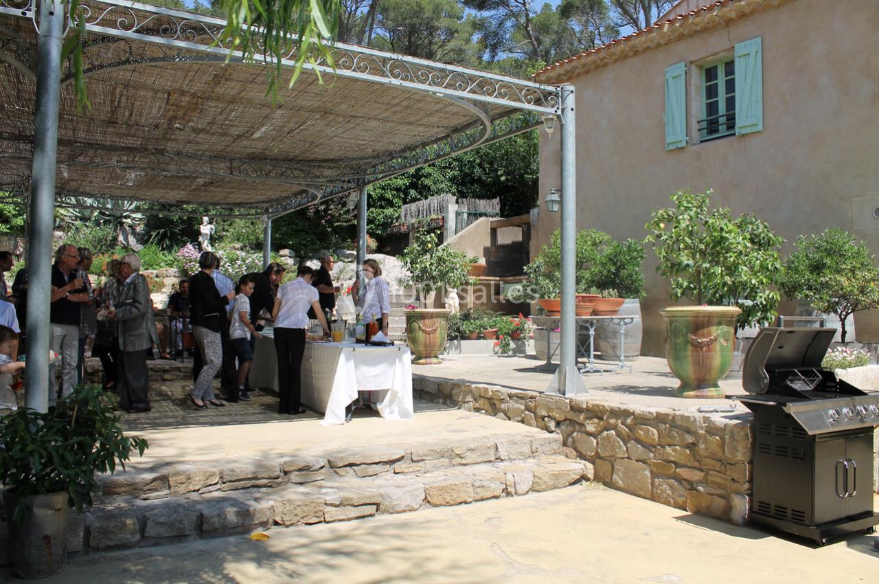 Terrasse ombragée avec des invités rassemblés autour d'une table, barbecue et plantes en pot à proximité.