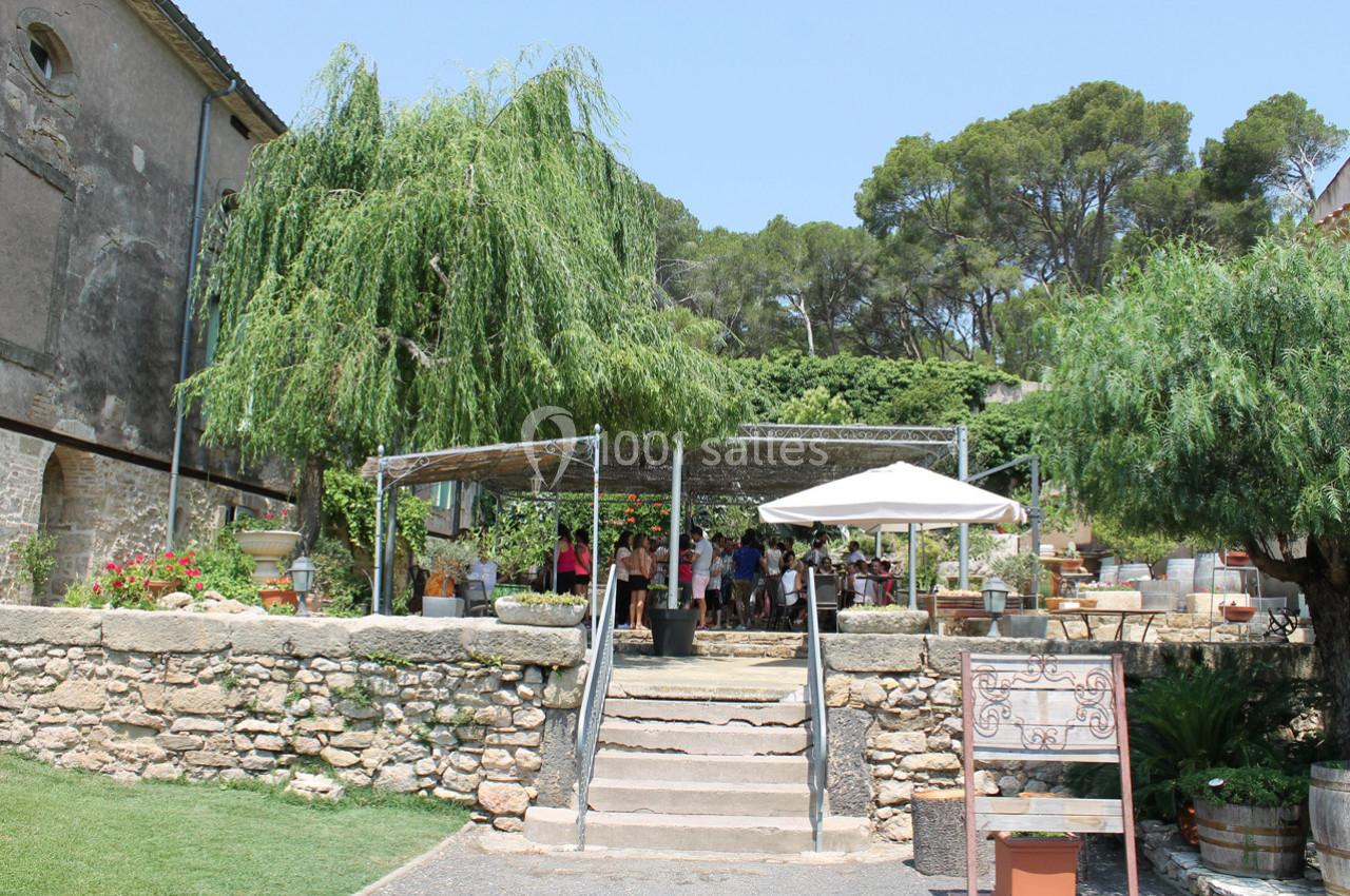 Terrasse ombragée avec des tables et des personnes, entourée de végétation et d'un bâtiment ancien en arrière-plan.
