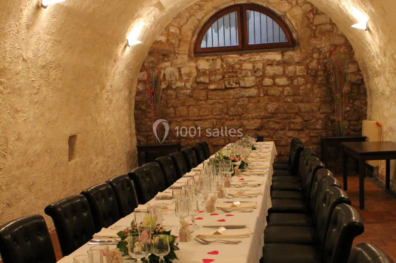 Salle voûtée en pierre avec une longue table dressée pour un repas, décorée de fleurs et de pétales rouges.