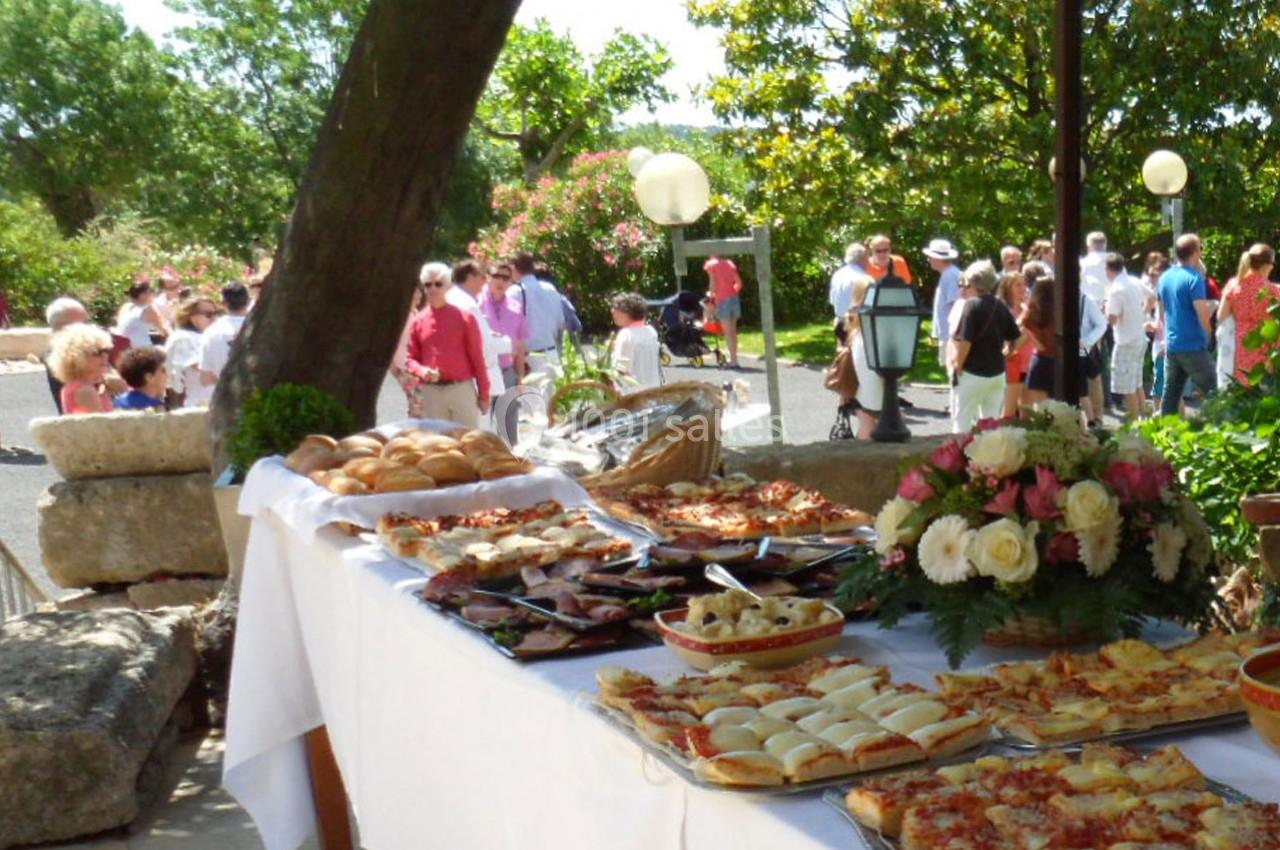 Buffet en plein air avec pizzas, pains et fleurs, entouré de convives dans un jardin ensoleillé.