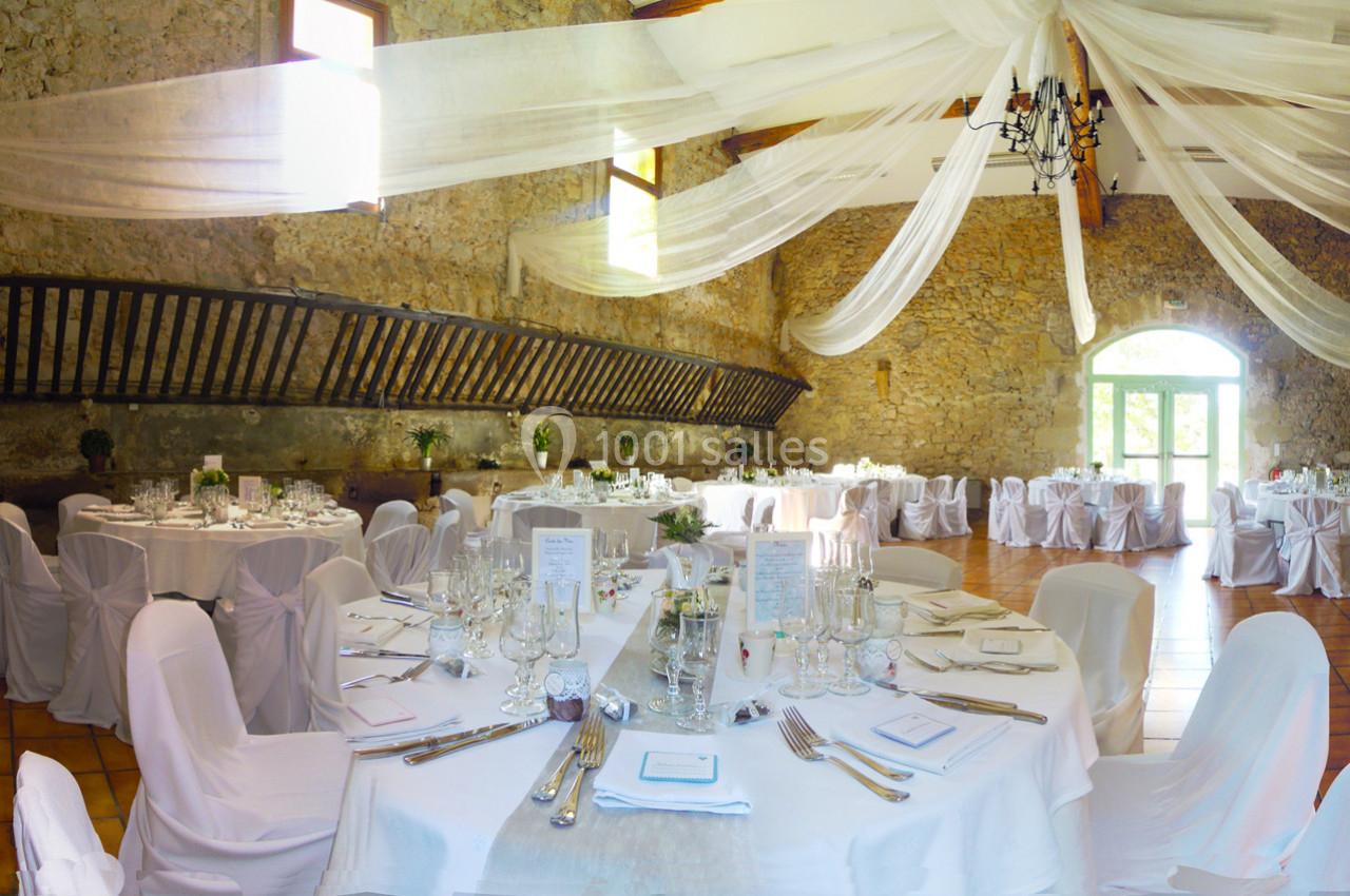 Salle de réception décorée pour un mariage, avec tables rondes dressées, chaises blanches et voilages suspendus.
