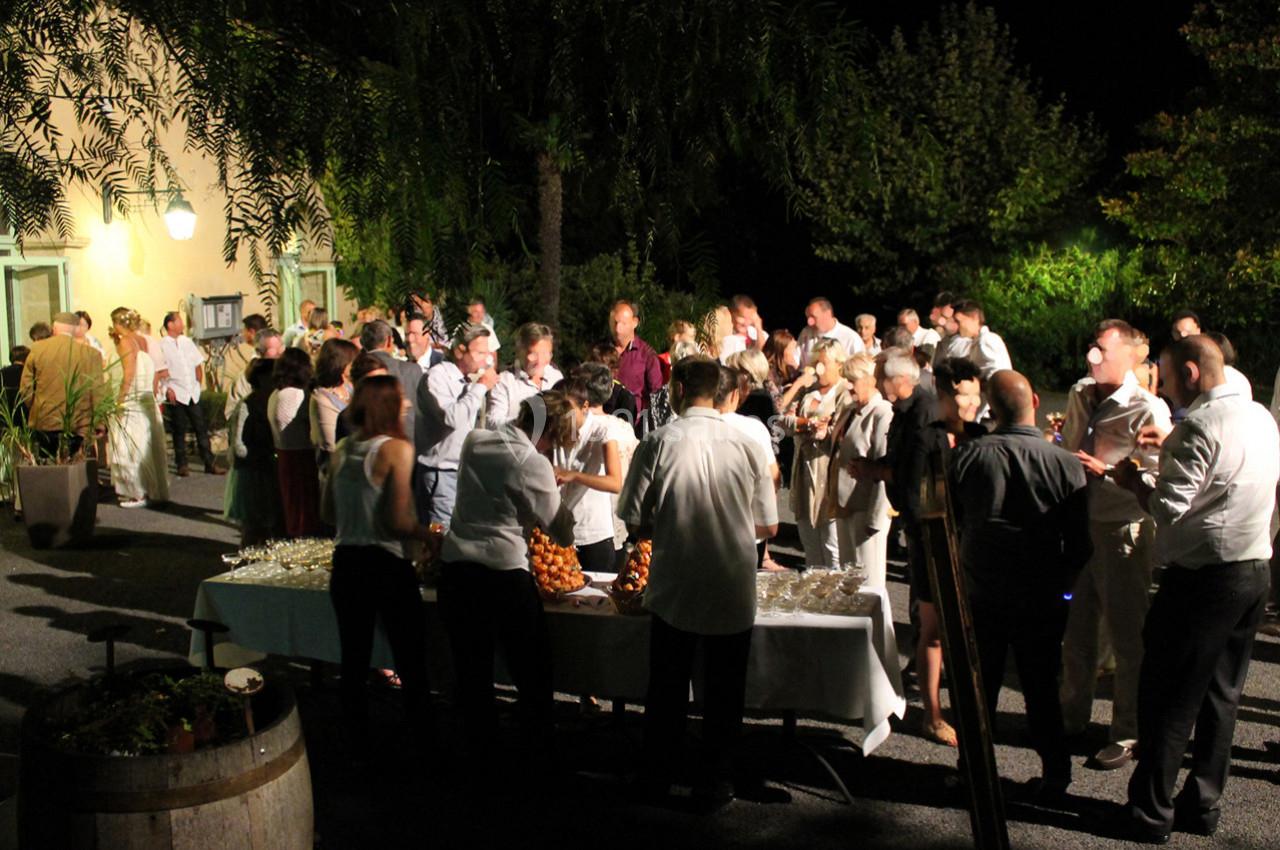 Groupe de personnes rassemblées en soirée autour d'une table de buffet en extérieur, éclairée par des lumières.