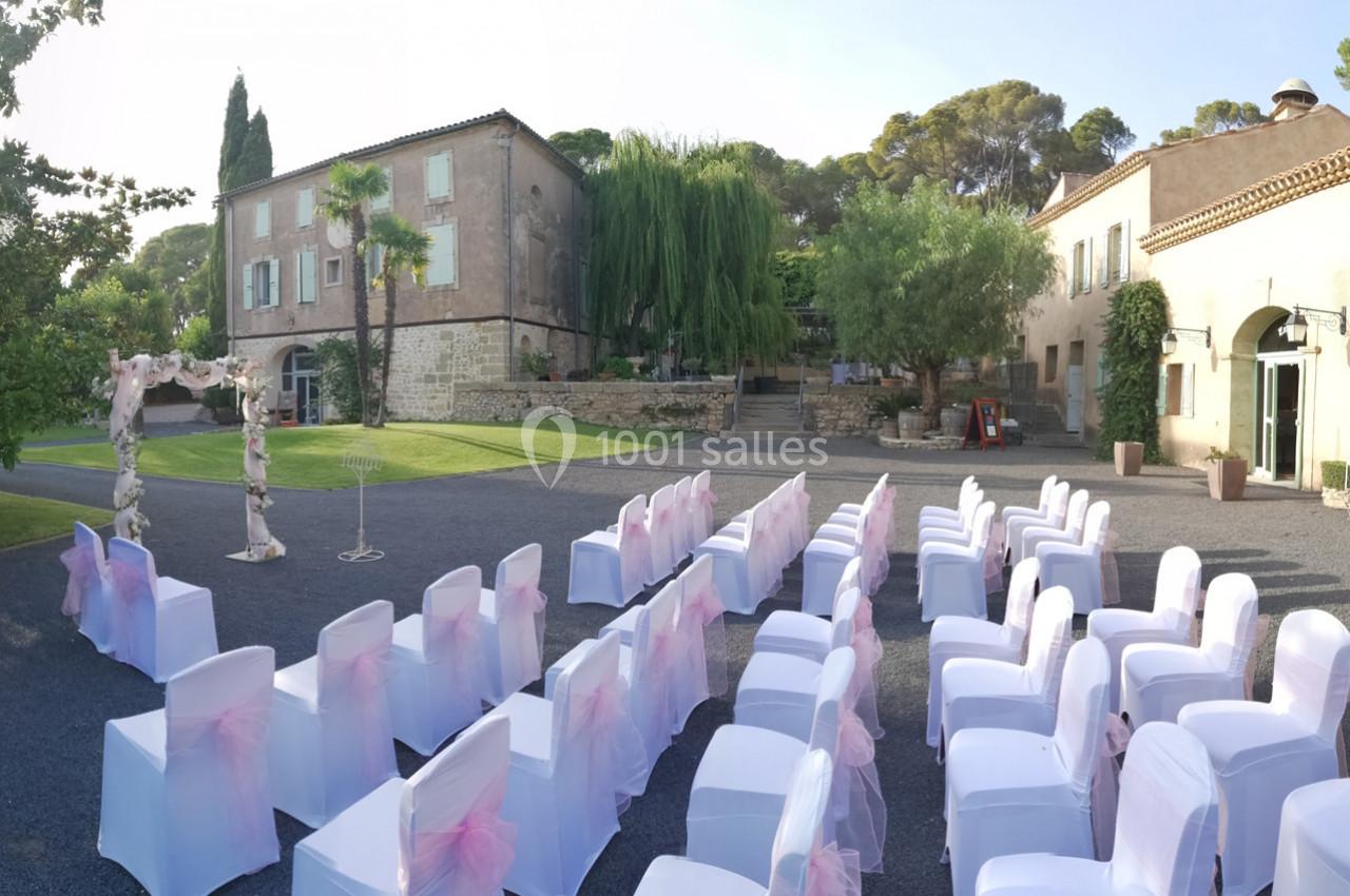 Chaises blanches décorées de rubans roses disposées en extérieur face à une arche florale, devant un bâtiment en pierre.