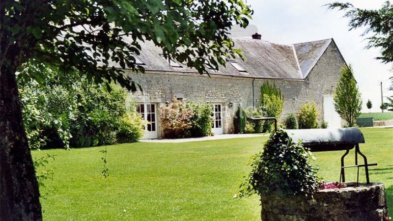 Maison en pierre entourée d'un jardin verdoyant avec un puits en premier plan et des arbres en arrière-plan.