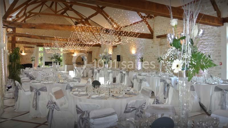 Salle de réception décorée avec des tables rondes, nappes blanches, chaises ornées de rubans argentés et guirlandes…
