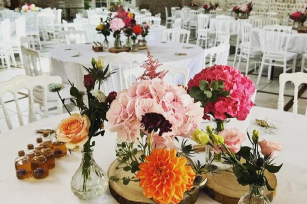 Salle de réception avec tables rondes décorées de bouquets colorés et petits pots en verre sur des rondins de bois.