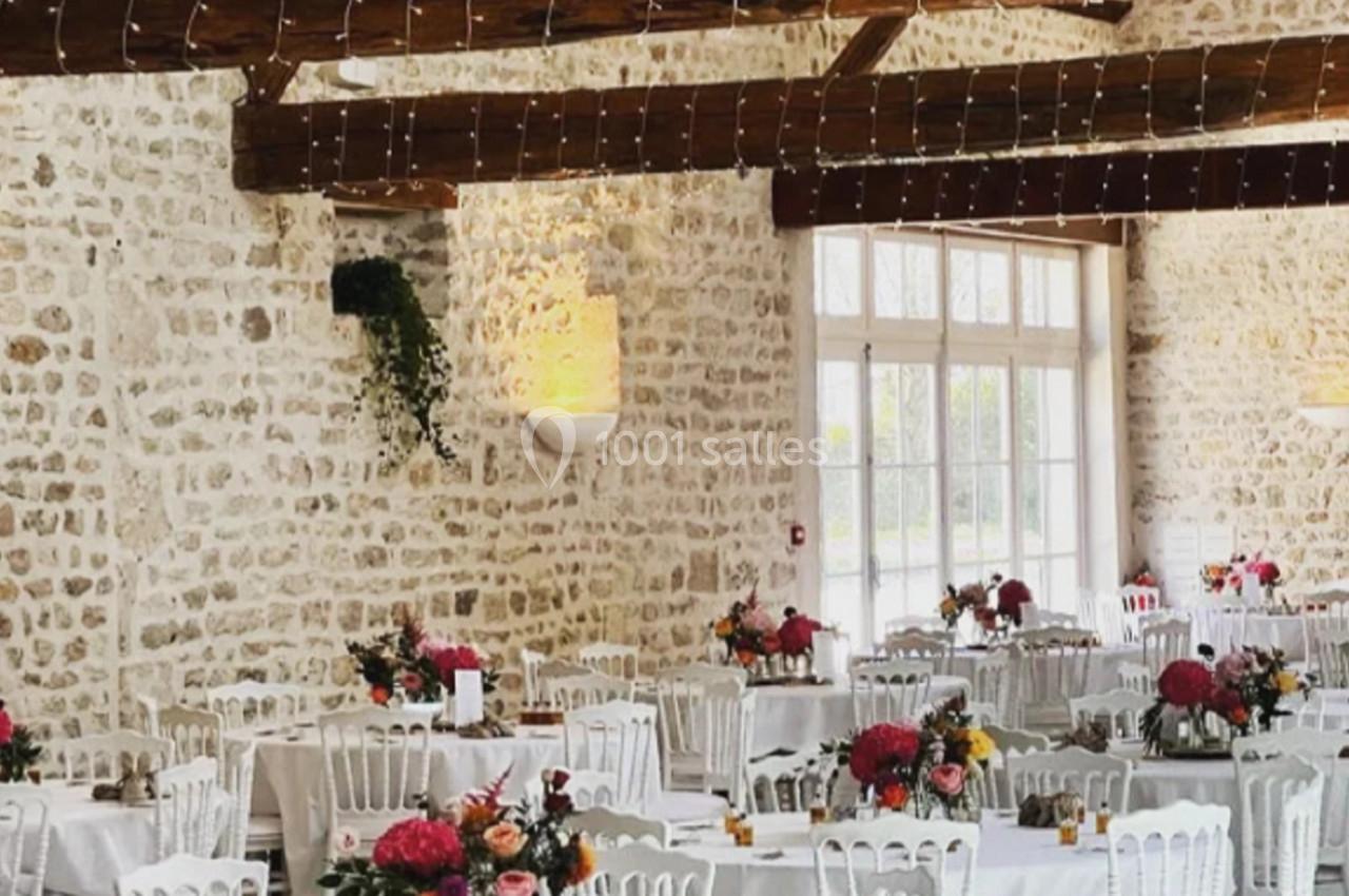 Salle de réception avec murs en pierre, tables rondes décorées de fleurs et chaises blanches sous des poutres apparentes.