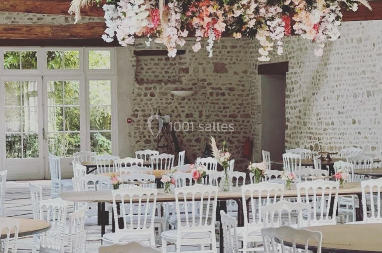Salle de réception lumineuse avec tables rondes, chaises blanches et une décoration florale suspendue au plafond.