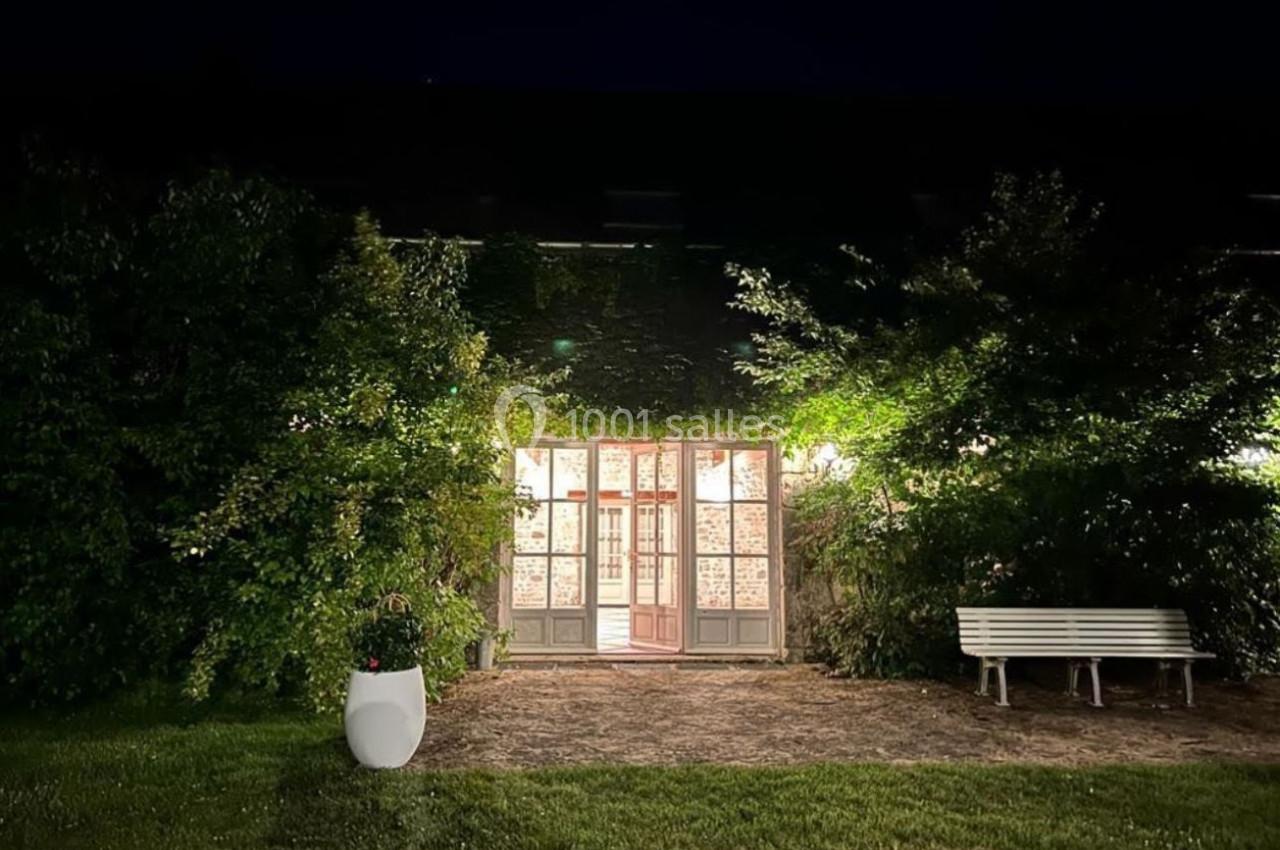 Façade vitrée éclairée entourée de végétation, avec un banc blanc et un pot de fleurs au premier plan, de nuit.