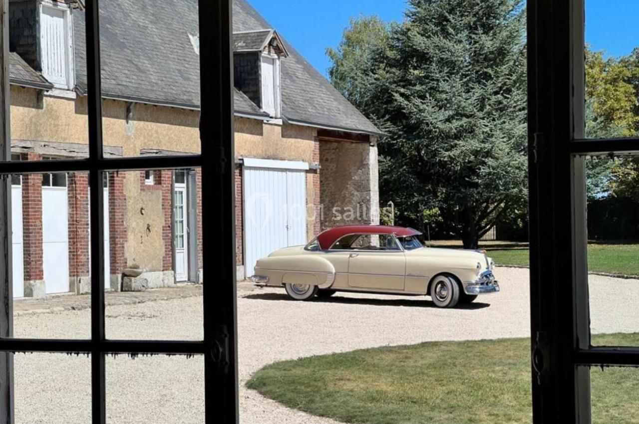 Voiture ancienne beige avec toit rouge garée dans une cour devant une maison en pierre et briques, vue depuis une fenêtre.