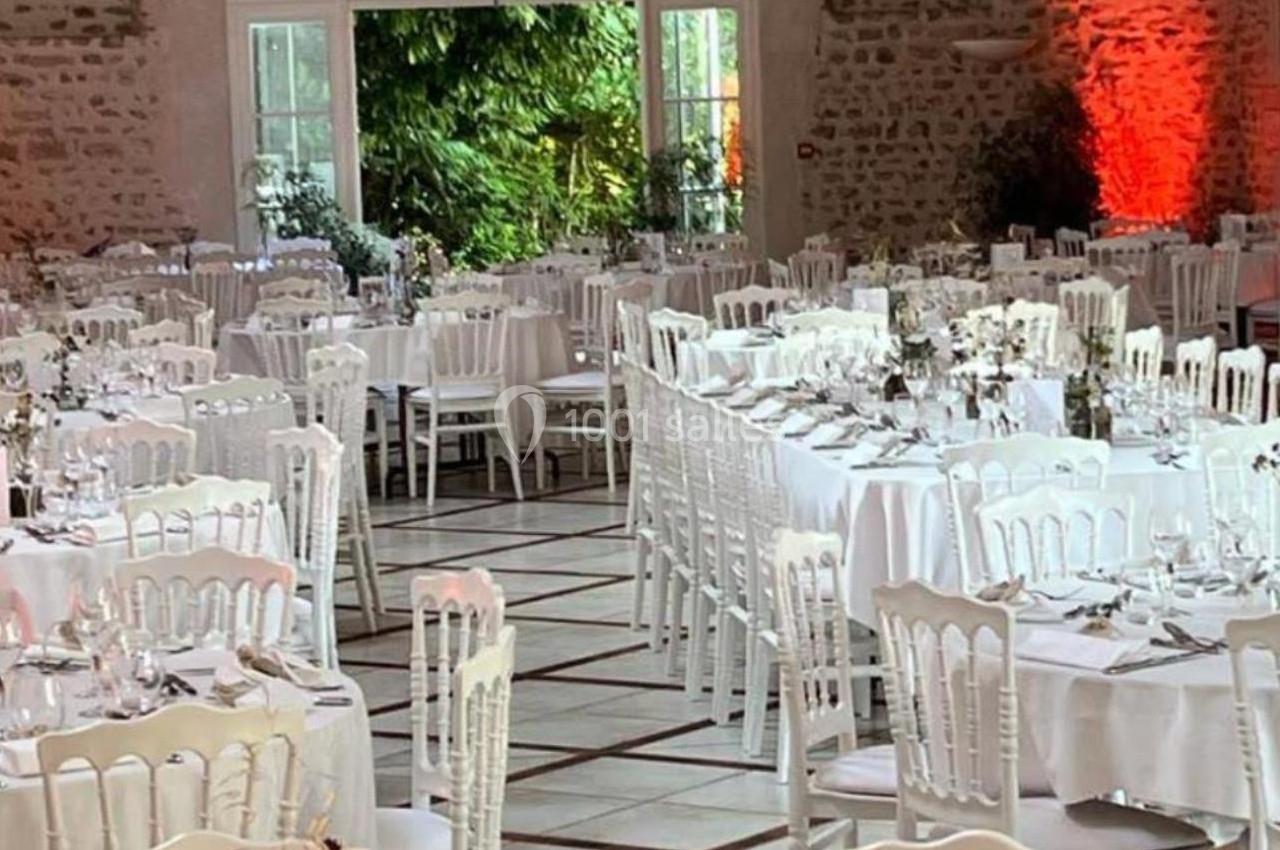 Salle de réception décorée avec des tables rondes dressées, chaises blanches et guirlandes lumineuses suspendues.