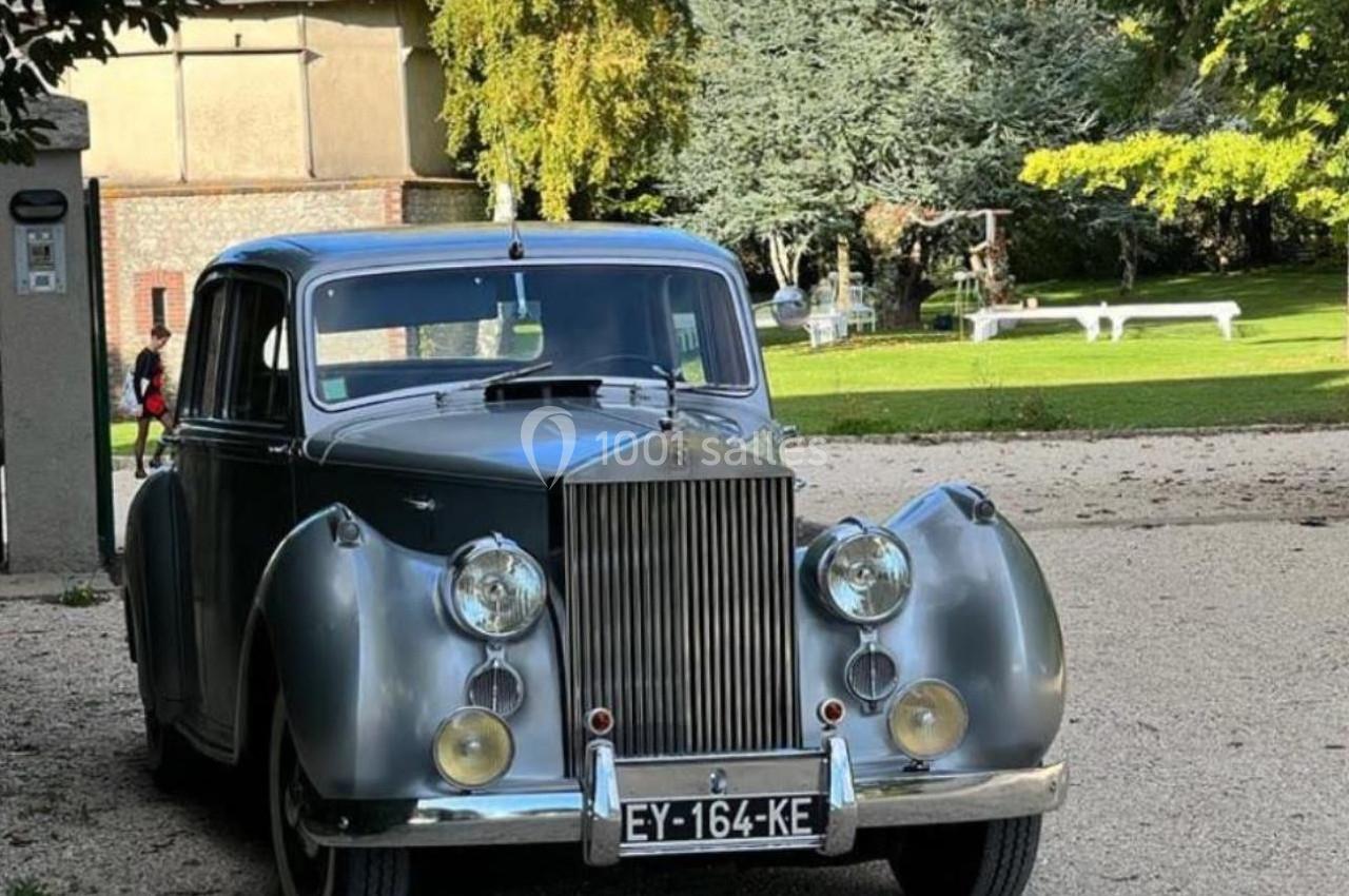 Voiture ancienne grise stationnée sur un chemin gravillonné, entourée d'arbres et d'un bâtiment en arrière-plan.
