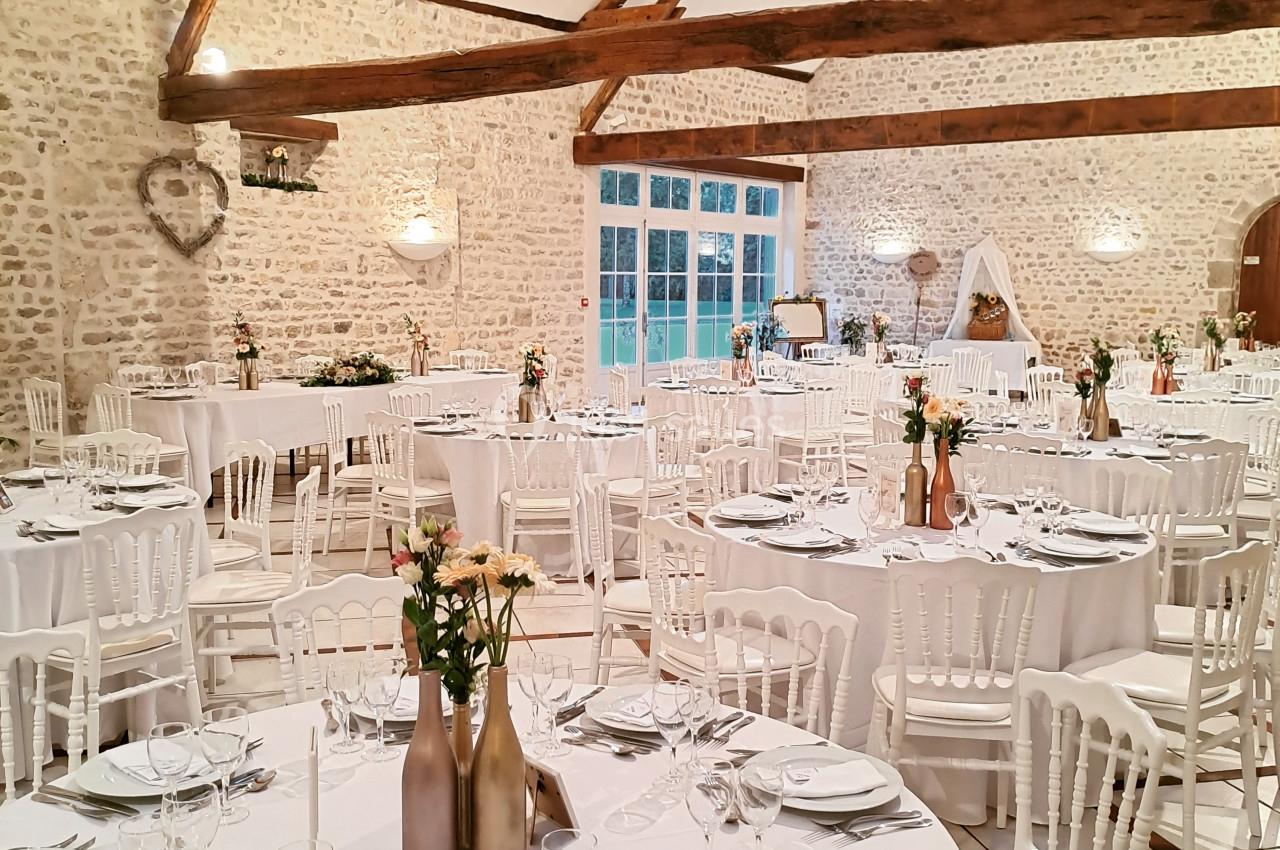 Salle de réception lumineuse avec murs en pierre, poutres apparentes et tables dressées pour un événement.