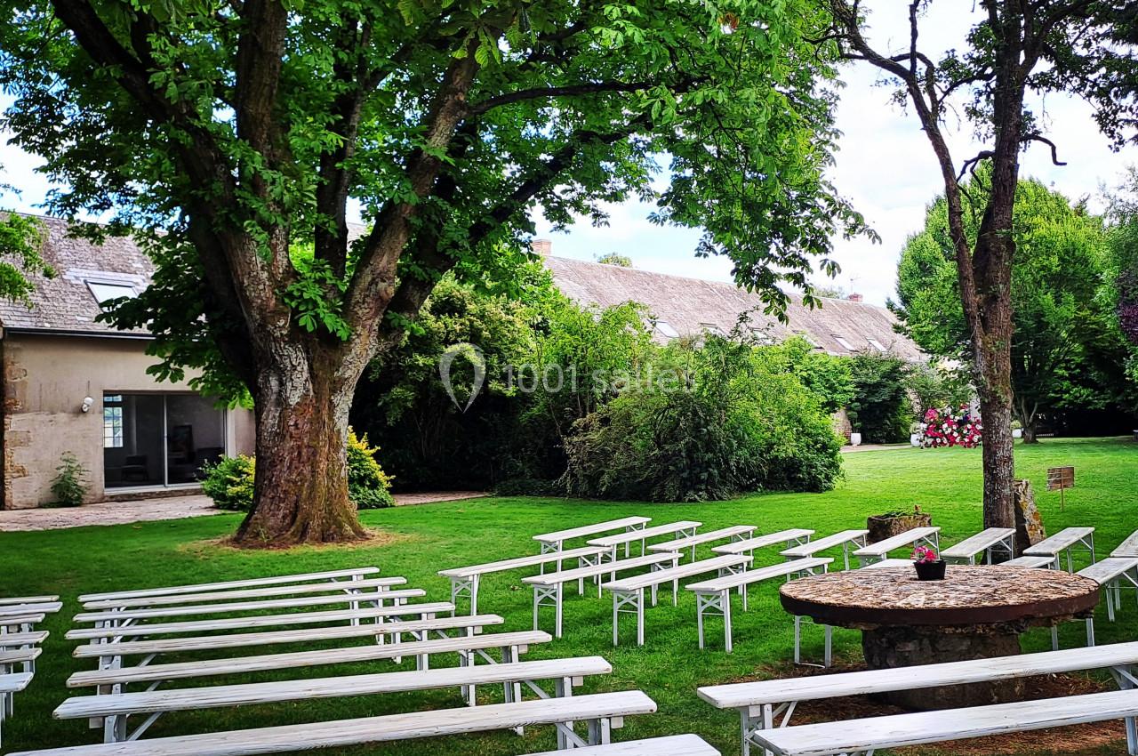 Bancs blancs alignés en plein air sous de grands arbres, face à une table ronde en bois dans un jardin verdoyant.