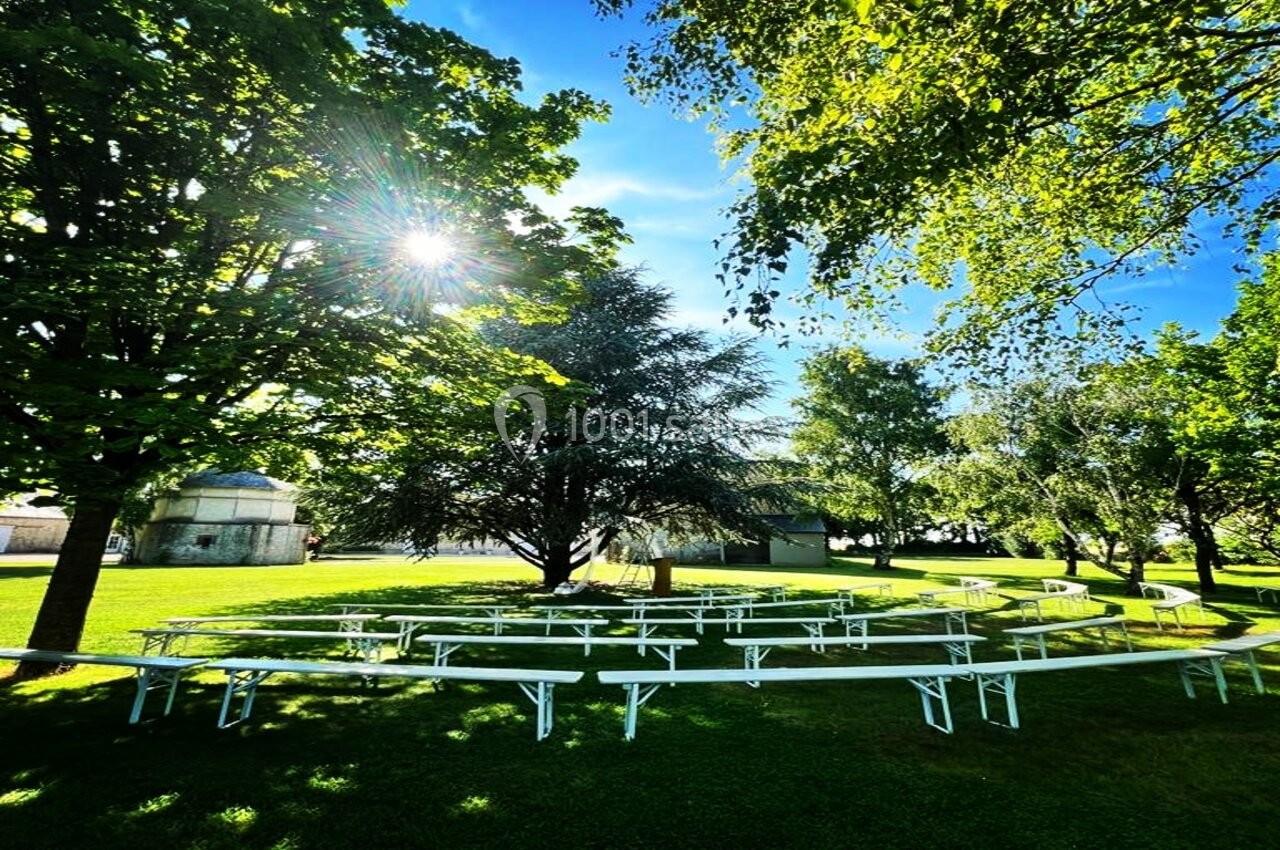 Tables et bancs blancs disposés sur une pelouse ombragée, entourés d'arbres sous un ciel ensoleillé.