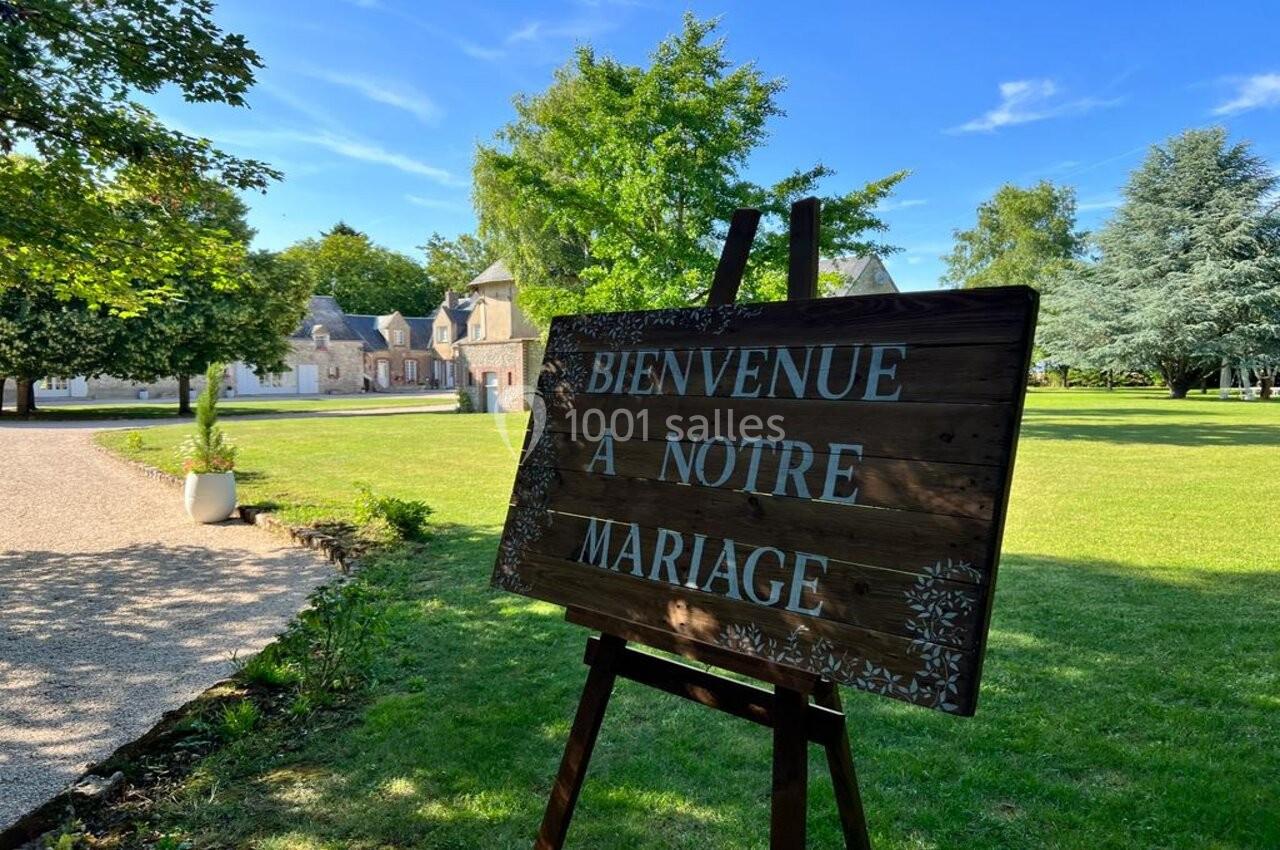 Panneau en bois ’Bienvenue à notre mariage’ sur un chevalet, devant un parc verdoyant et un bâtiment en arrière-plan.