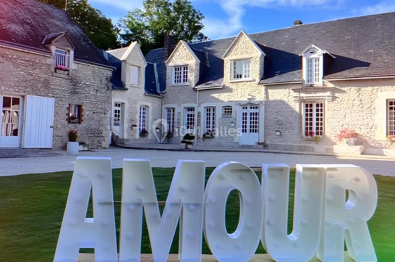 Grandes lettres blanches formant le mot ’AMOUR’ devant une maison en pierre avec des volets et un toit en ardoise.