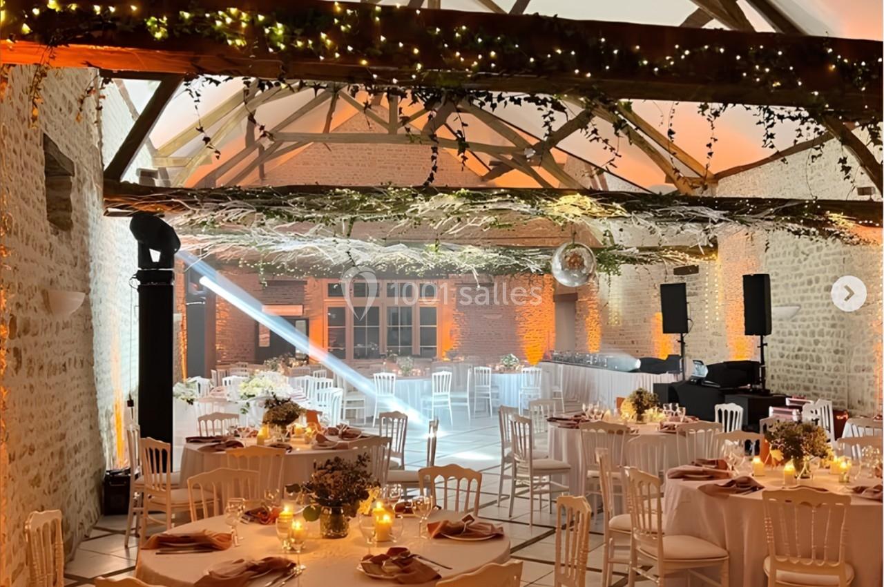 Salle de réception décorée avec des tables rondes, des chaises blanches, des lumières chaleureuses et des poutres apparentes.