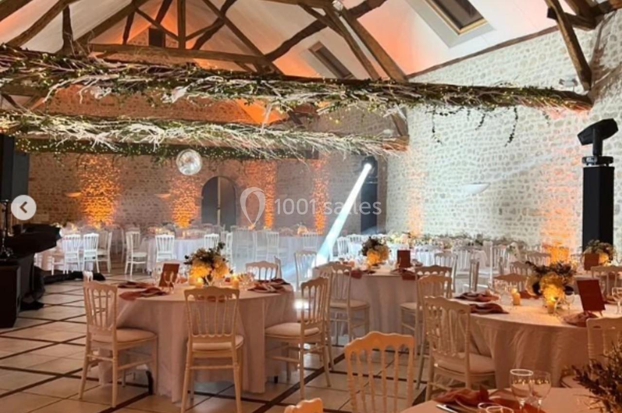 Salle de réception décorée avec des tables rondes, nappes blanches, guirlandes lumineuses et poutres apparentes.