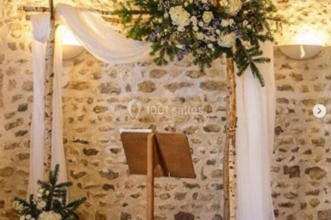 Arche de mariage en bois décorée de fleurs blanches et vertes, installée devant un mur en pierre.