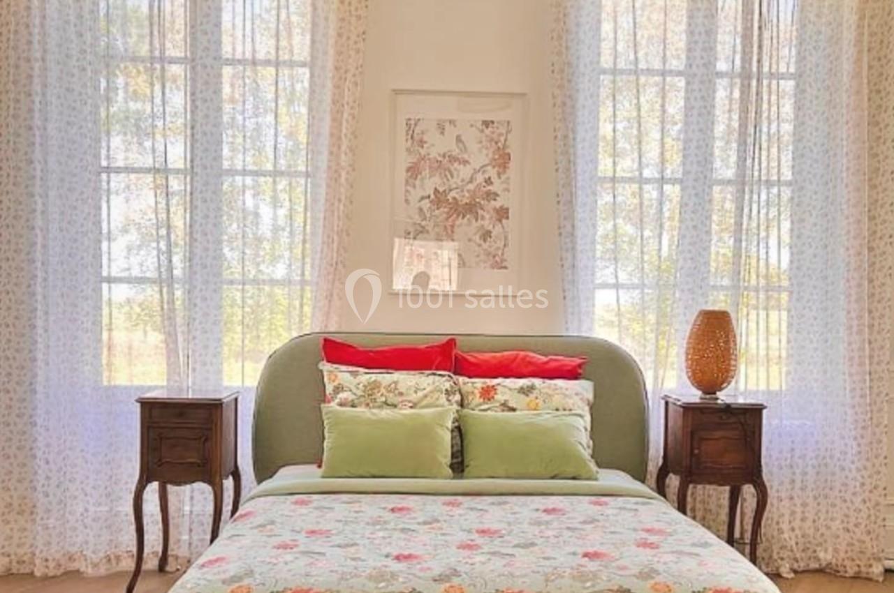 Chambre lumineuse avec lit à motifs floraux, tables de chevet en bois et grandes fenêtres encadrées de rideaux légers.