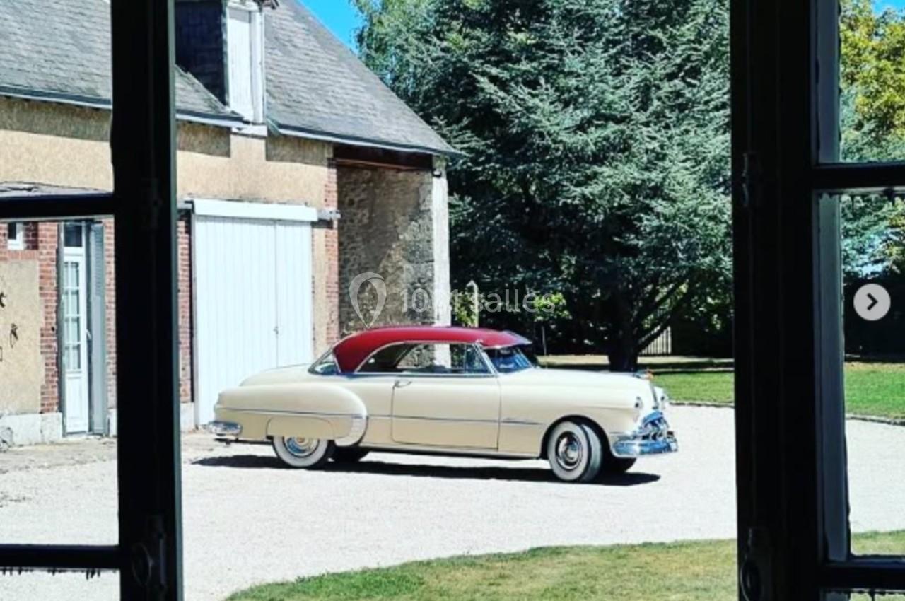 Voiture ancienne beige et rouge garée dans une cour gravillonnée, vue depuis l'intérieur d'une maison.