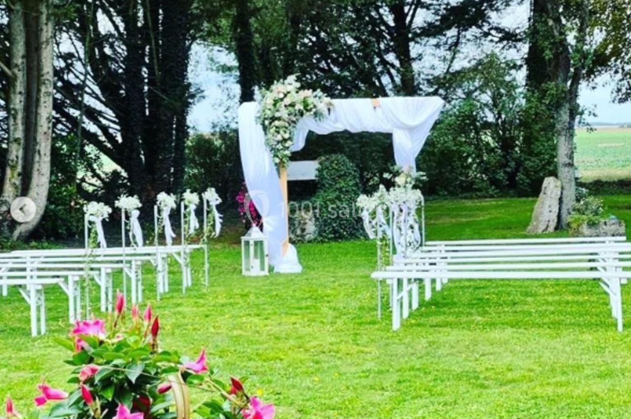Arche de mariage décorée de fleurs blanches, entourée de bancs blancs, dans un jardin verdoyant.